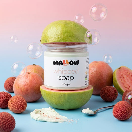 A jar of MALLOW whipped soap stands centered on a blue surface with fruit props around it, including lychees, and guavas. Bubbles float in the background against a pink-to-blue gradient backdrop.