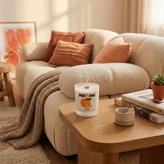A frosted glass jar peach candle labeled 'MALLOW' sits on a wooden side table in a cozy living room setting with a cream boucle sofa, rust-orange pillows, a knitted throw blanket, and books nearby.