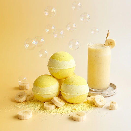 Three round yellow bath bombs with pale cream centers are arranged on a light beige surface with scattered banana slices and powder. A glass of pale yellow drink with a banana slice on the rim sits beside them, with floating bubbles in the background.