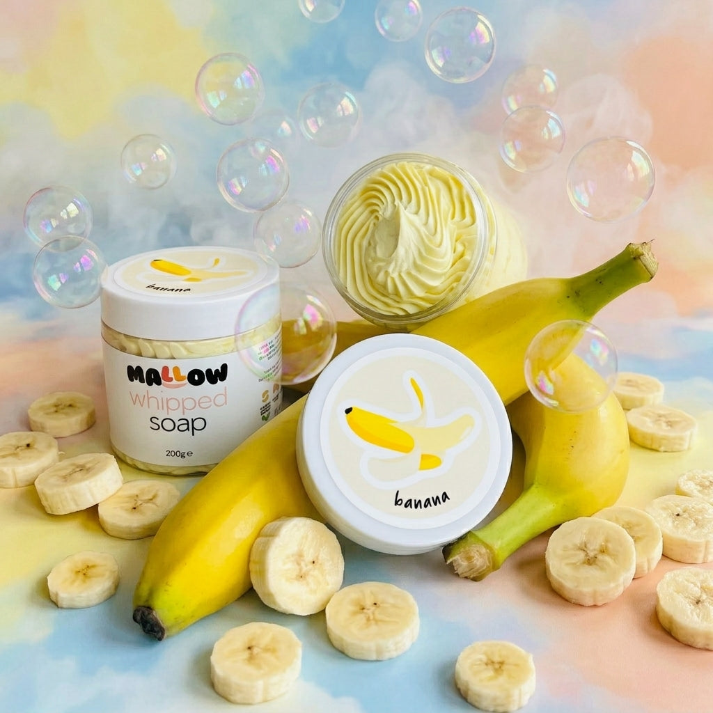 Mallow Whipped Soap in banana scent, shown with a jar, an open tub revealing whipped yellow soap, and a closed container with a banana illustration label. The products are styled with fresh bananas, sliced bananas, and floating bubbles on a pastel cloud background.