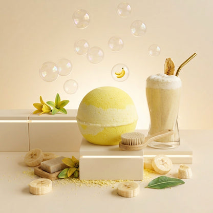 A styled bath-and-body product scene on a warm cream background. A round yellow bath bomb sits on a pedestal next to a frothy milkshake in a glass with a gold straw, a wooden brush, banana slices, soap bars, green leaves, and floating bubbles.