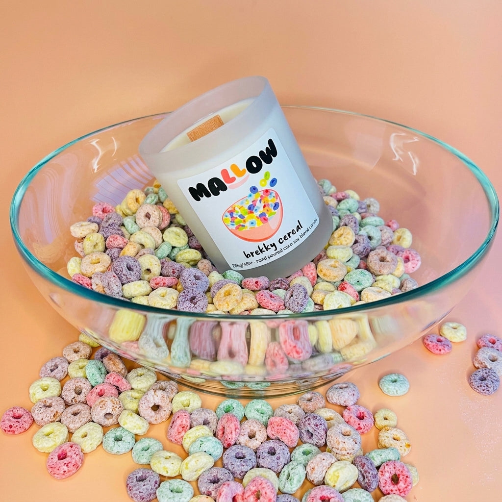 A frosted glass jar candle labeled 'MALLOW brekky cereal' sits in a large clear glass bowl filled with pastel multicolour cereal pieces, with more cereal scattered around on a peach-pink background.