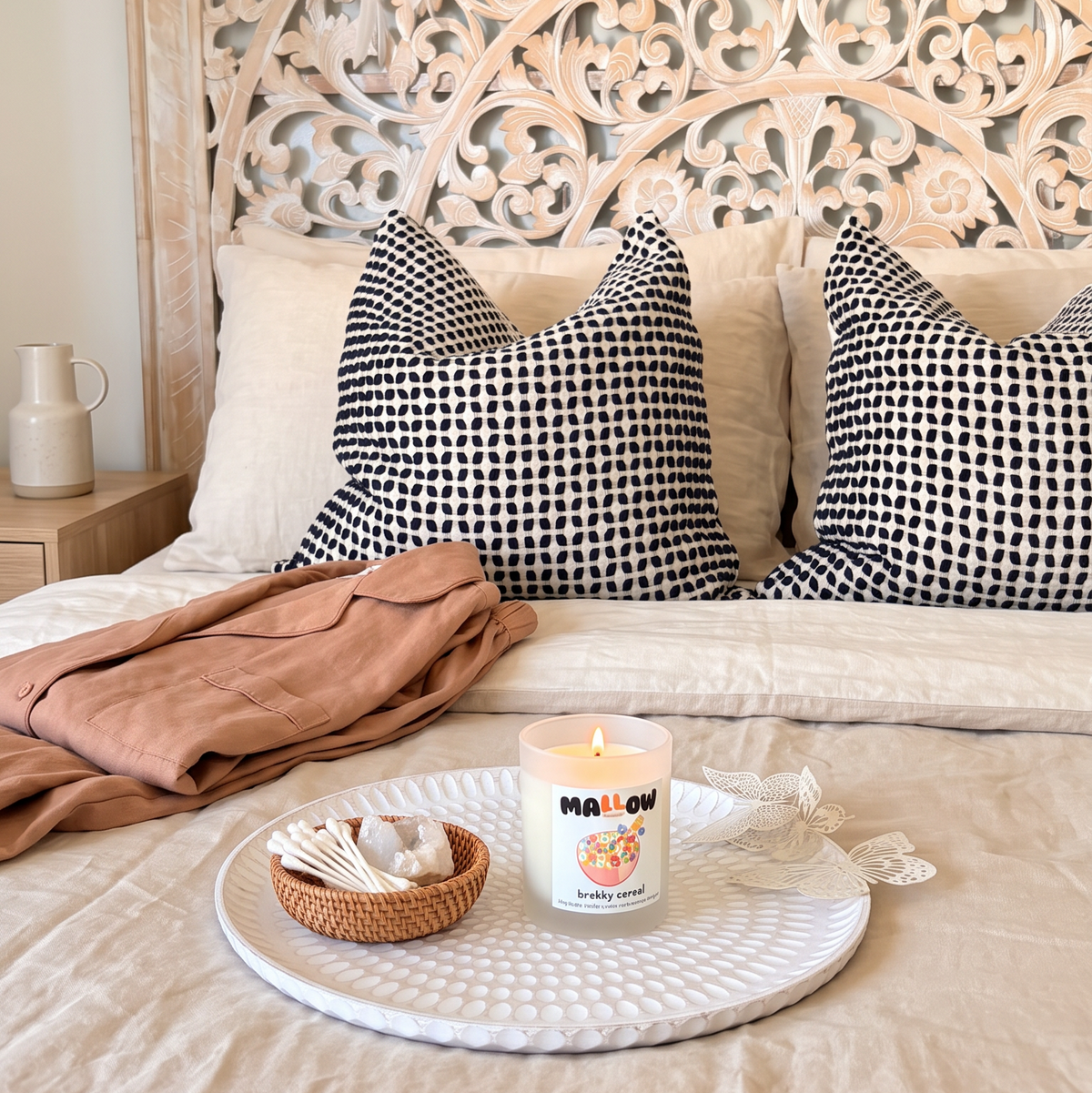 A styled bedroom scene with a bed dressed in beige linens, two black-and-white patterned pillows, a folded tan throw, and a tray holding a lit white candle labeled 'Mali Light' in front of an ornate carved wooden headboard.