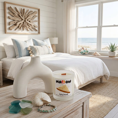 A frosted glass jar fairy bread candle with a white sculptural base in a bright beach-style bedroom. The candle label 'MALLOW'.