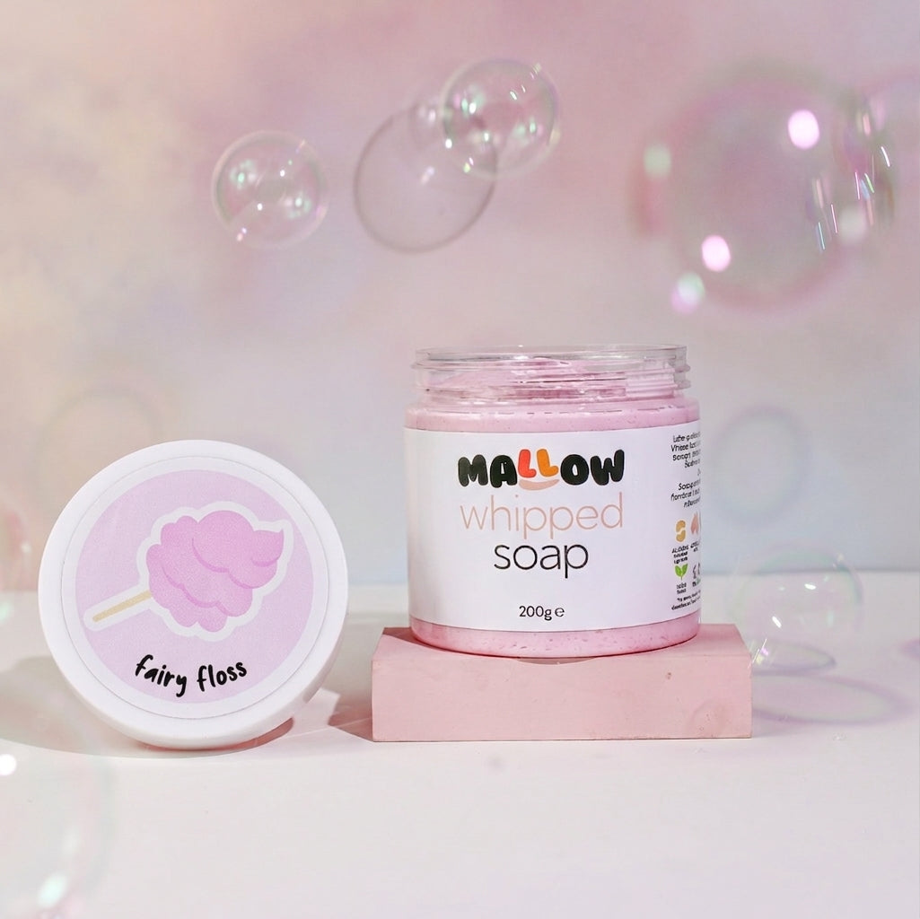 A jar of Mallow Whipped Soap and its matching lid are displayed on a pink block against a soft pink background with floating bubbles. The lid features a pink cotton candy illustration labeled 'Fairy Floss'.