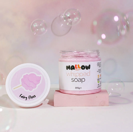 A jar of Mallow Whipped Soap and its matching lid are displayed on a pink block against a soft pink background with floating bubbles. The lid features a pink cotton candy illustration labeled 'Fairy Floss'.