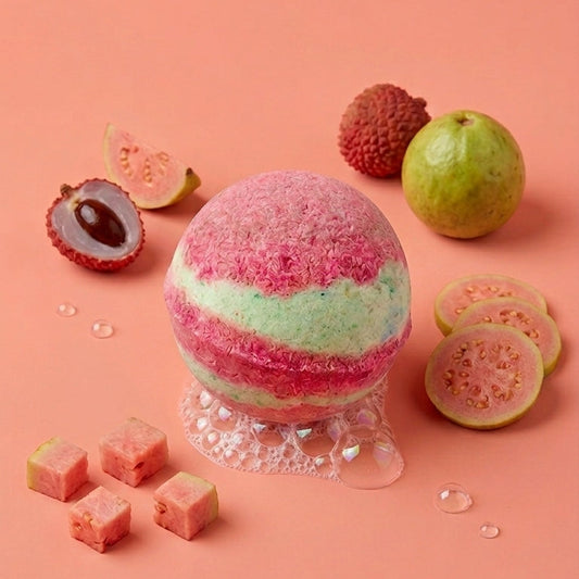 A spherical bath bomb with pink and mint green layers sits on a clear pedestal, surrounded by sliced guava, whole guava, and pink fruit cubes on a solid peach-pink background.