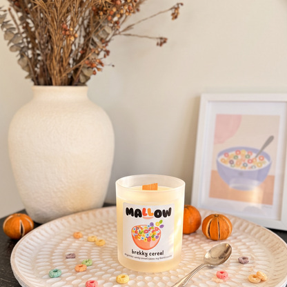 A Mallow brand candle styled on a textured white tray with a spoon, scattered cereal pieces, small oranges, and dried flowers in a vase in the background.