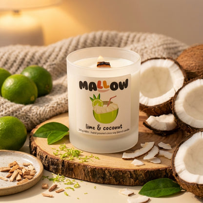 A frosted glass candle labeled 'MALLOW lime & coconut' sits on a rustic wood slice surrounded by limes, coconut halves, coconut flakes, and a knitted beige cloth. The candle is lit with a warm glow, and the background is softly blurred.