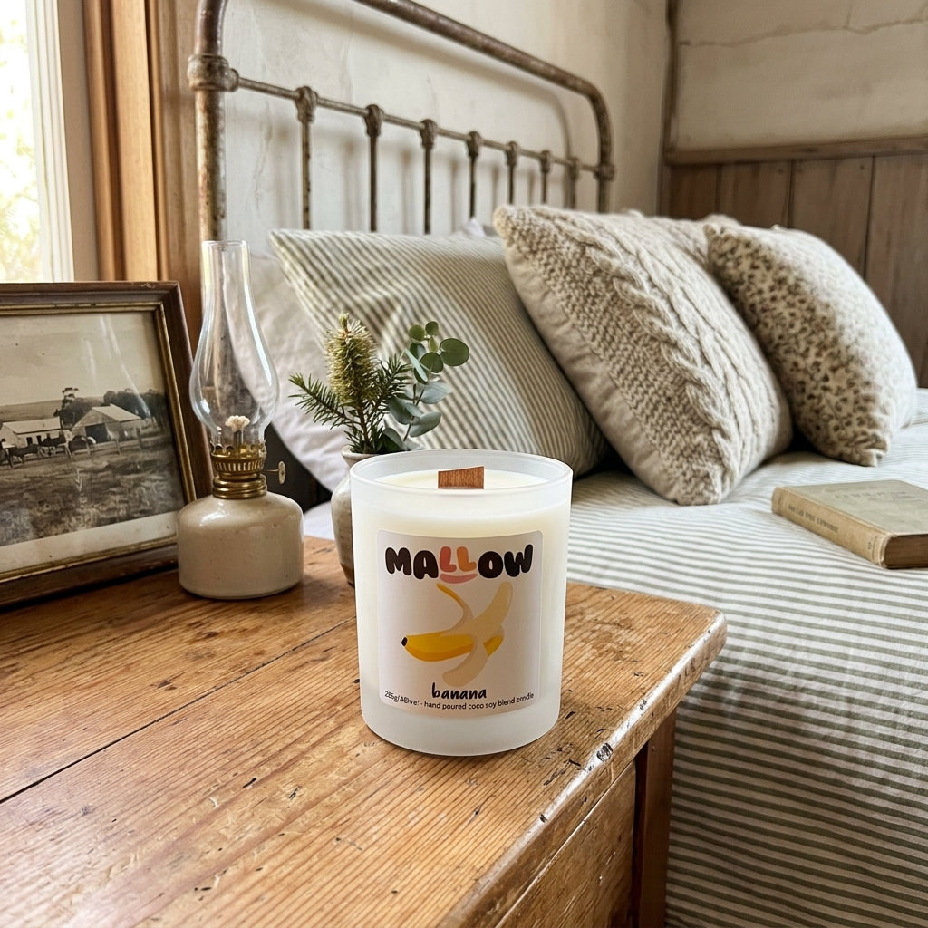 A frosted glass jar candle labeled 'MALLOW banana' sits on a rustic wooden bedside table in a cozy bedroom setting with a metal bed frame, striped bedding, and textured pillows.