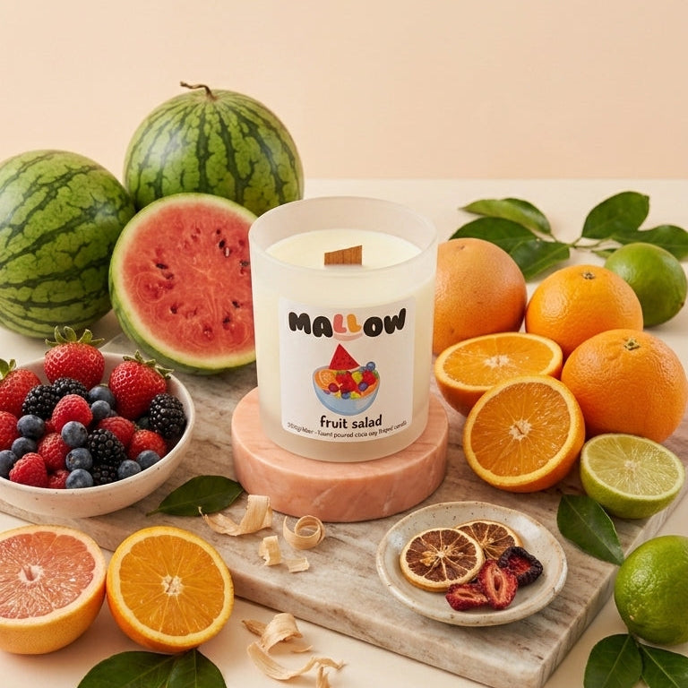 A fruit-scented candle by the brand Mallow, styled on a wooden board with fresh fruit including watermelons, oranges, limes, and berries against a warm beige background.
