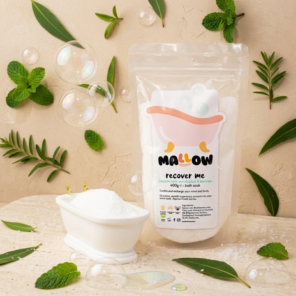 A stand-up pouch of MALLOW Recover Me bath soak stands on a light surface beside a small white tub filled with white powder, surrounded by mint leaves, green sprigs, bubbles, and scattered bath salts against a warm beige textured background.