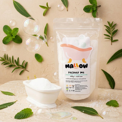 A stand-up pouch of MALLOW Recover Me bath soak stands on a light surface beside a small white tub filled with white powder, surrounded by mint leaves, green sprigs, bubbles, and scattered bath salts against a warm beige textured background.