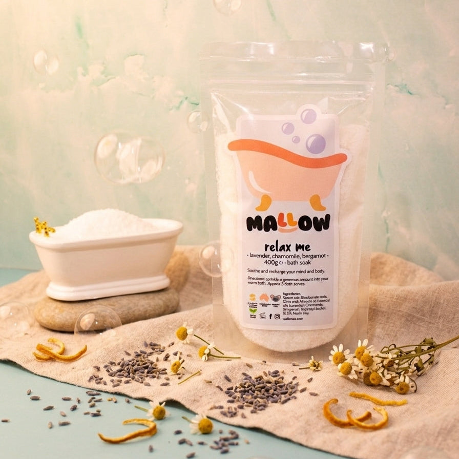 A stand-up pouch of Mallow Relax Me bath salts sits on a beige cloth with dried chamomile flowers, lavender buds, and orange peel scattered around it. A small white tub filled with white bath salts is to the left, set against a soft pastel background with floating bubbles.