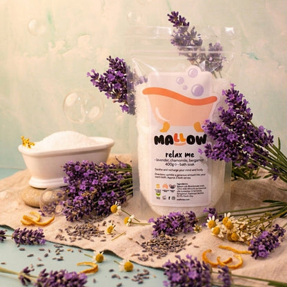 A clear stand-up pouch labeled 'MALLOW relax me' with a cartoon bath bubble graphic, styled with lavender sprigs, dried flowers, and a small white bowl on a beige cloth against a pale mint background.