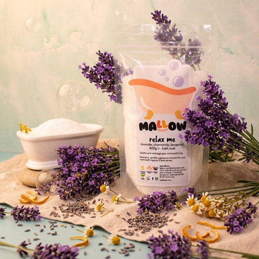 A clear stand-up pouch labeled 'MALLOW relax me' with a cartoon bath bubble graphic, styled with lavender sprigs, dried flowers, and a small white bowl on a beige cloth against a pale mint background.