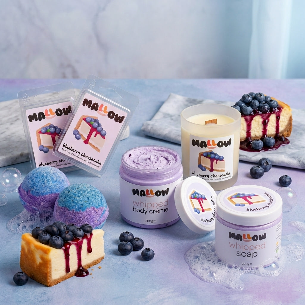 A styled product display of MALLOW brand blueberry cheesecake including scented wax melt, whipped body creams, a candle, and a whipped soap, bath bomb, arranged with blueberries and a cheesecake slice on a pastel blue surface.