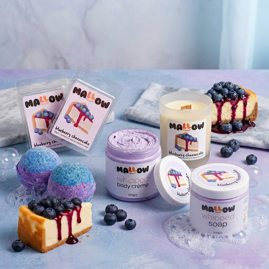 A styled product display of MALLOW brand blueberry cheesecake including scented wax melt, whipped body creams, a candle, and a whipped soap, bath bomb, arranged with blueberries and a cheesecake slice on a pastel blue surface.